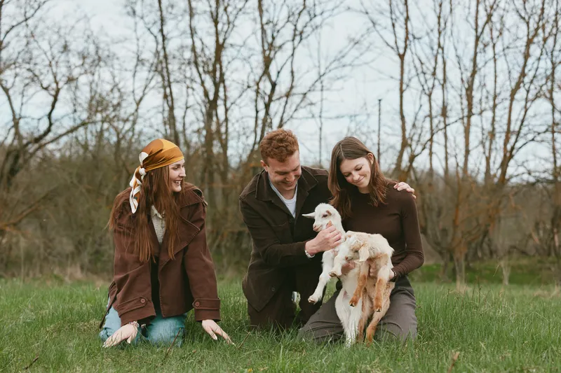 Cheerful group outdoors with a young goat kid, friends sharing a gentle moment on lush grass, candid interaction, natural light, relaxed mood, countryside setting