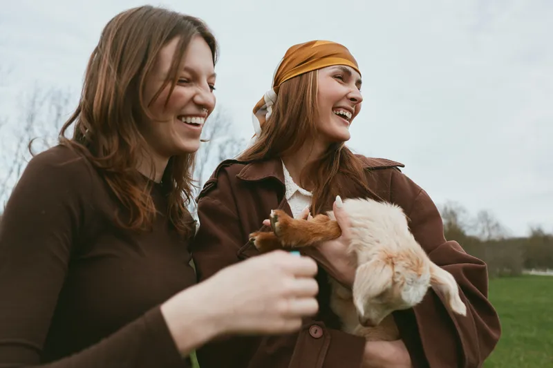 Two young women enjoy a candid moment outdoors while holding a baby goat, smiling and dressed in casual autumn clothing with natural background and soft daylight.