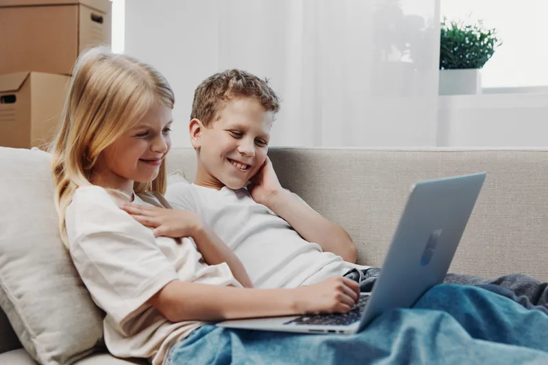 Two smiling children sitting on sofa using laptop together in bright room, enjoying technology and leisure time at home.