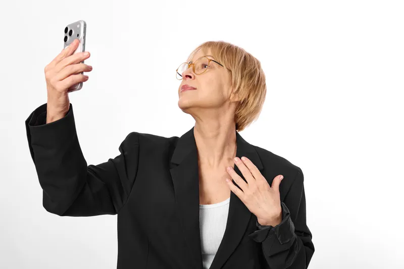 Elderly woman with glasses in business jacket takes a selfie using a smartphone on a white studio background neutral thoughtful pose confident expression and raised hand