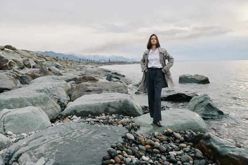 Woman standing confidently on rocky shore near sea wearing coat and trousers. Overcast sky with distant mountains visible. Concept of confidence, nature, and solitude outdoors.