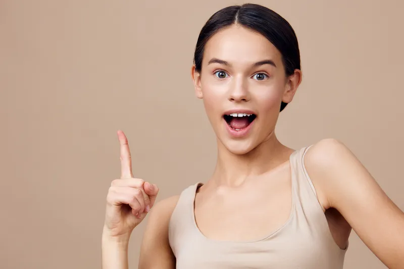 Joyful young woman celebrating a creative idea with a finger raised, wearing a beige top against a warm neutral background, exuding positivity and inspiration