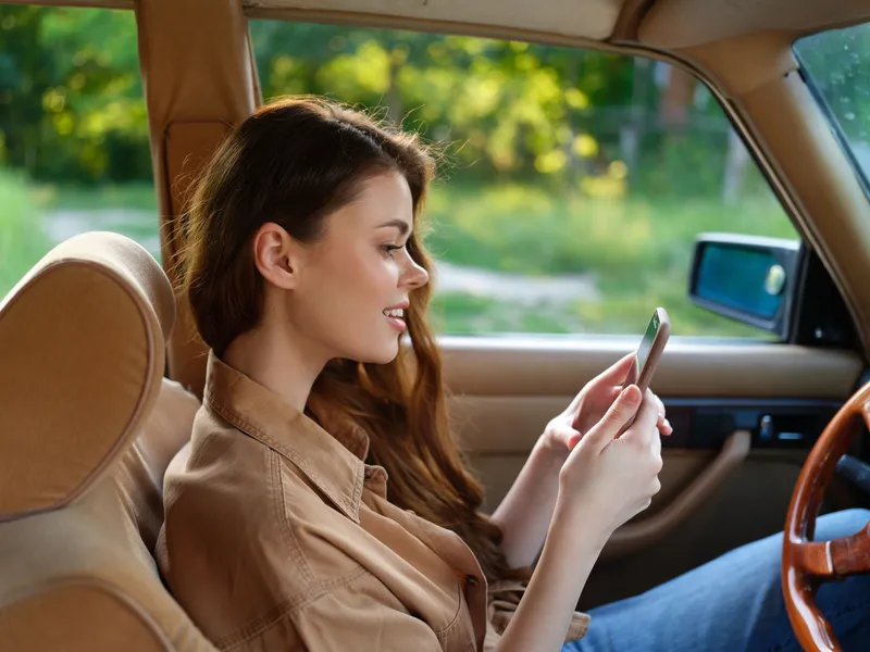 Young woman in casual outfit sitting in classic car, smiling while using smartphone, surrounded by natural greenery and soft sunlight.