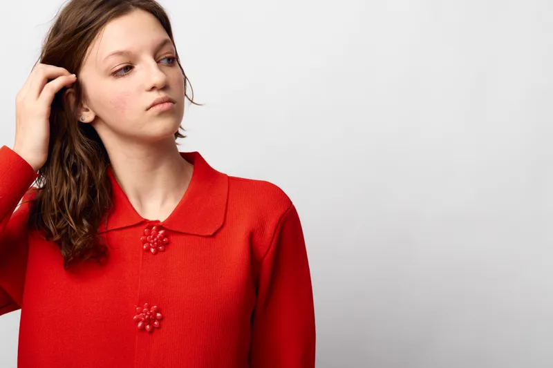 Young woman in red sweater looks away thoughtfully, touching her hair. Minimalist studio background with soft lighting. Concept of contemplation, fashion, and casual style.