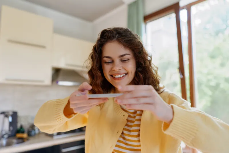 Young woman in a yellow sweater smiles while looking at her phone in a bright kitchen, capturing a casual, everyday home moment of connection and warmth.
