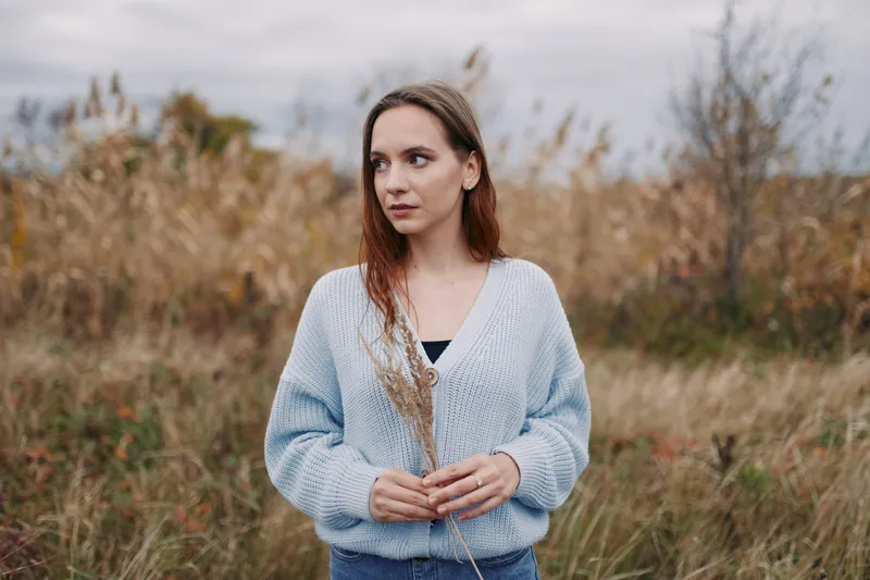 Portrait of a young woman standing in a windswept meadow, holding dried grasses, natural light and soft tones convey an authentic, honest mood and a simple, realistic moment.