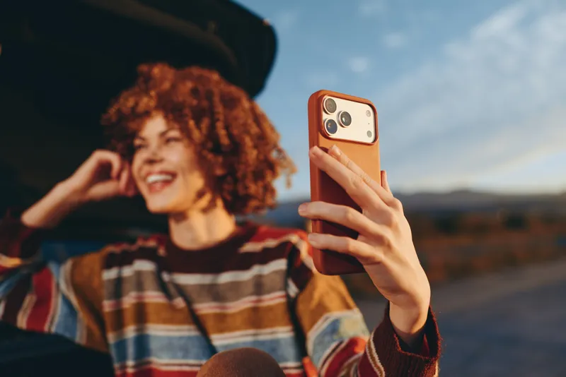 Woman lifestyle selfie with smartphone outdoors, smiling curly hair and hat in striped sweater at sunset, hand holding phone in foreground, softly blurred background travel scene