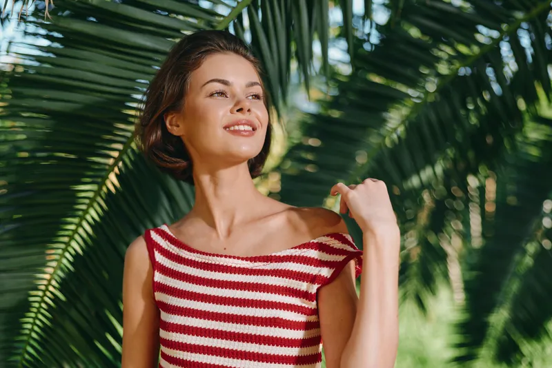 A cheerful young woman stands outdoors among lush palm leaves, wearing a striped red and white top. She smiles warmly, enjoying sunlight and a breezy, relaxed moment.