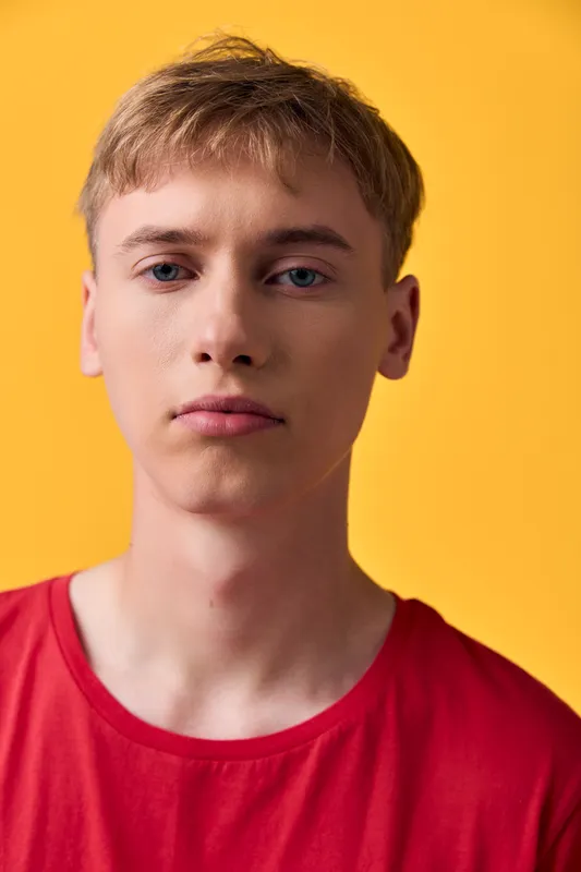 Young man with a calm expression against a bright yellow background wearing a red shirt in a close up shot with studio lighting and clean backdrop emphasizing color and mood