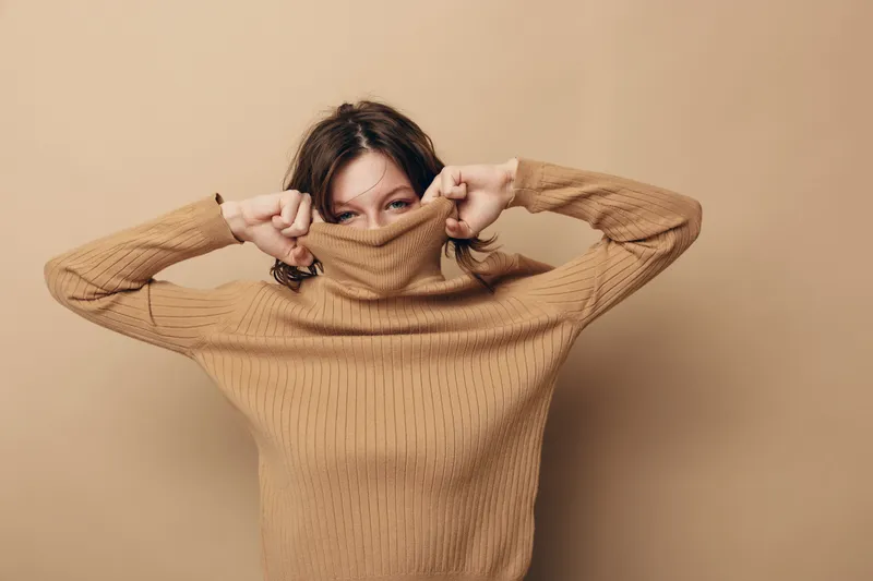 Young woman in beige knitted sweater pulls collar over face, peeking with eyes. Studio portrait on neutral background. Concept of cozy fashion, autumn style, and playful mood.