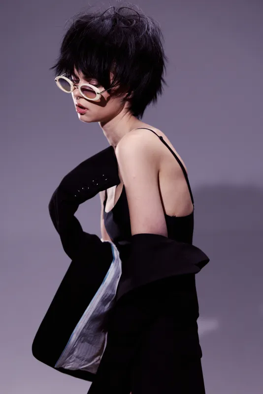 Stylish woman with short hair wearing sunglasses and black outfit in minimal purple background fashion pose.