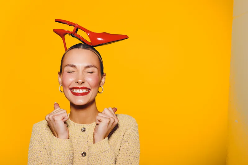 Happy woman with closed eyes smiles joyfully wearing red high heel shoe on her head against bright yellow background. Concept of fun, fashion, and playful mood in studio portrait.