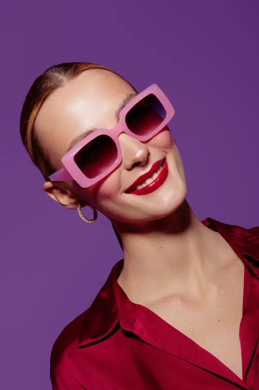 Smiling woman with pink sunglasses and red lipstick poses against purple background. She wears hoop earrings and a red blouse. Concept of fashion, beauty, and summer style.