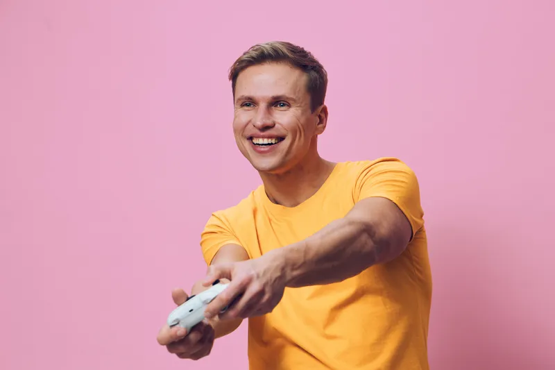 smiling young man in orange t-shirt hold gamepad play video game isolated on pink background digital entertainment concept