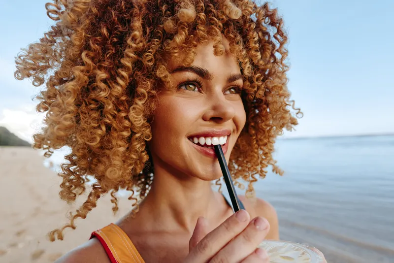 Young woman with curly hair enjoys a cold drink on a sunny beach while smiling and looking into the distance with a relaxed and happy expression.