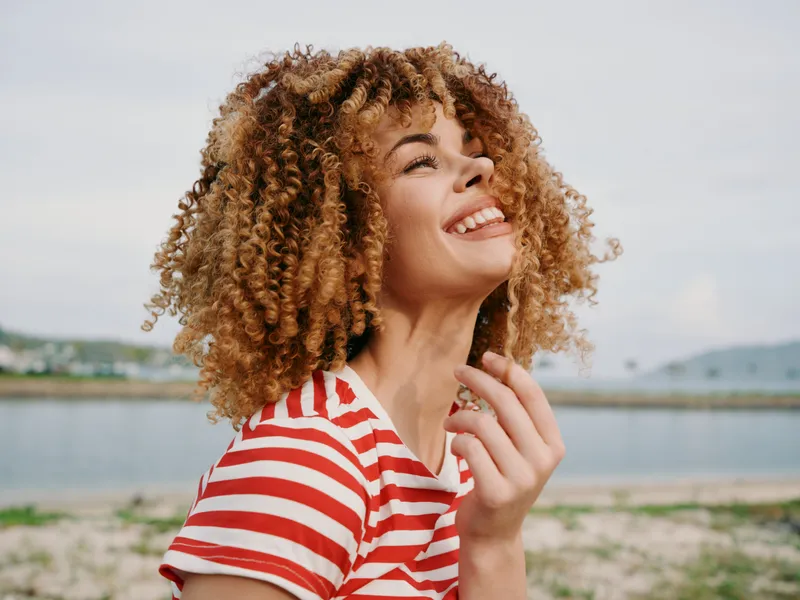 Joyful portrait of a woman with curly hair in a red and white striped shirt, smiling by the seaside with wind in her hair and bright natural light.
