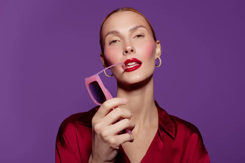 Stylish woman with red lipstick and hoop earrings holds pink sunglasses near lips against purple background. Fashion portrait with confident expression and vibrant colors for beauty concept.