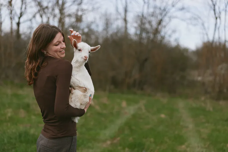 Woman outdoors carries a small kid goat in a grassy field, smiling warmly, showcasing rural lifestyle and bonding with animals, candid moment of countryside companionship under soft natural light