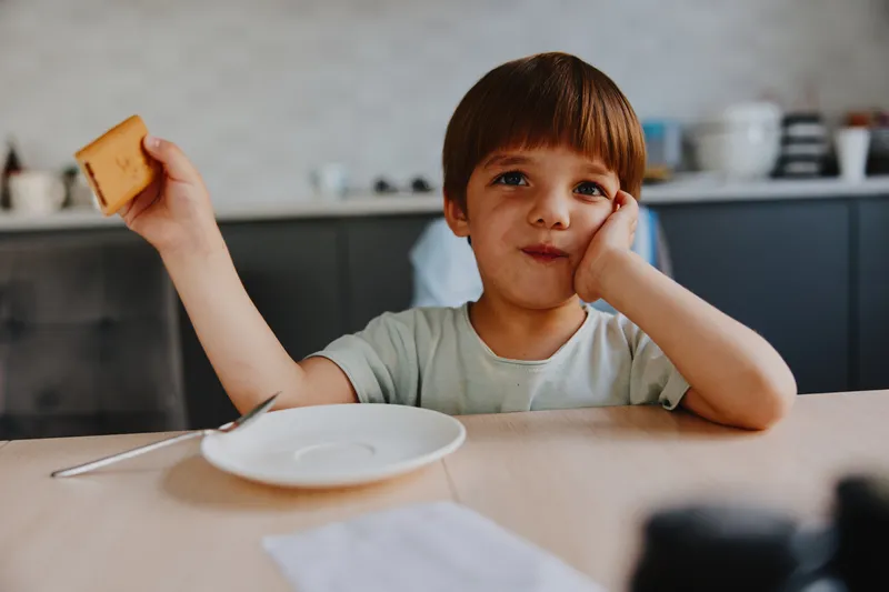 Thoughtful boy holding a piece of bread, sitting at the table. He looks curious and contemplative, with a focused expression in a cozy kitchen setting.