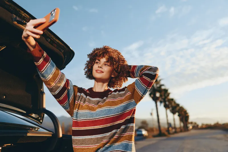 Woman lifestyle, outdoors by a car on a road at sunset, curly hair, striped sweater, holding smartphone up for a selfie, relaxed joyful pose, warm golden light, evening breeze, city trees
