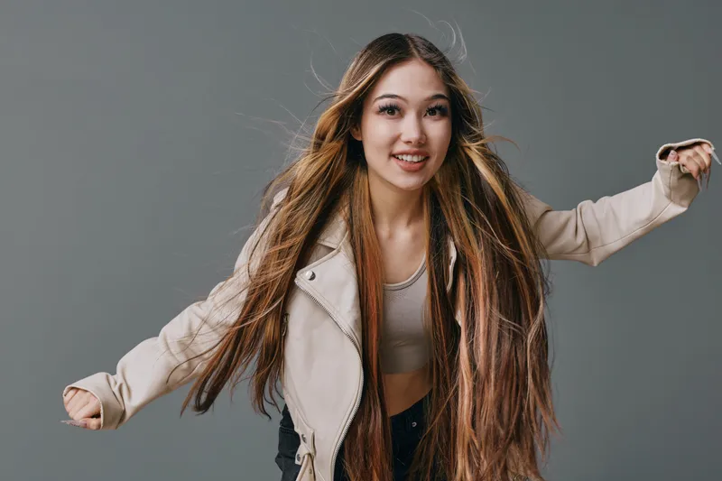 Young woman smiling and dancing with long straight hair and casual outfit. Stylish female model wearing jacket and crop top posing against grey background with joyful expression.