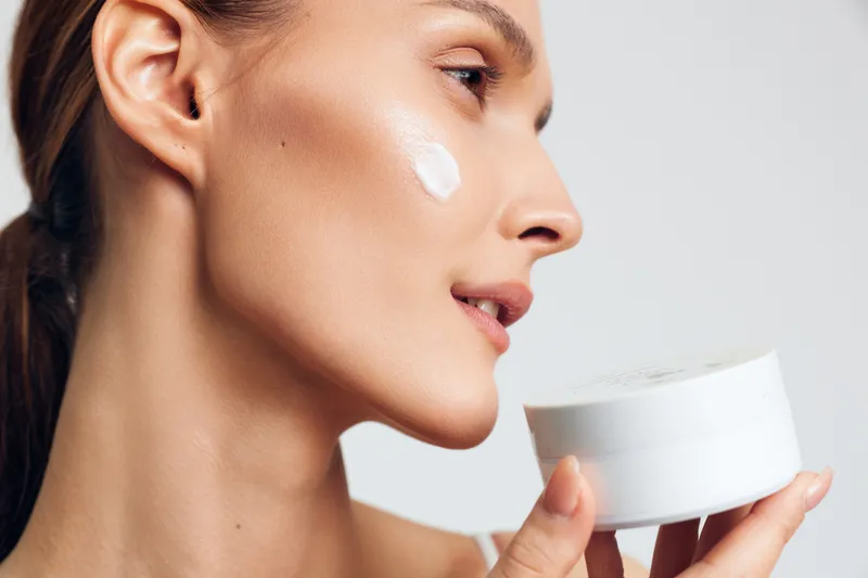Young woman applying white cream on cheek, skincare routine, closeup profile, natural makeup, smooth skin, soft lighting, beauty concept, wellness, personal care