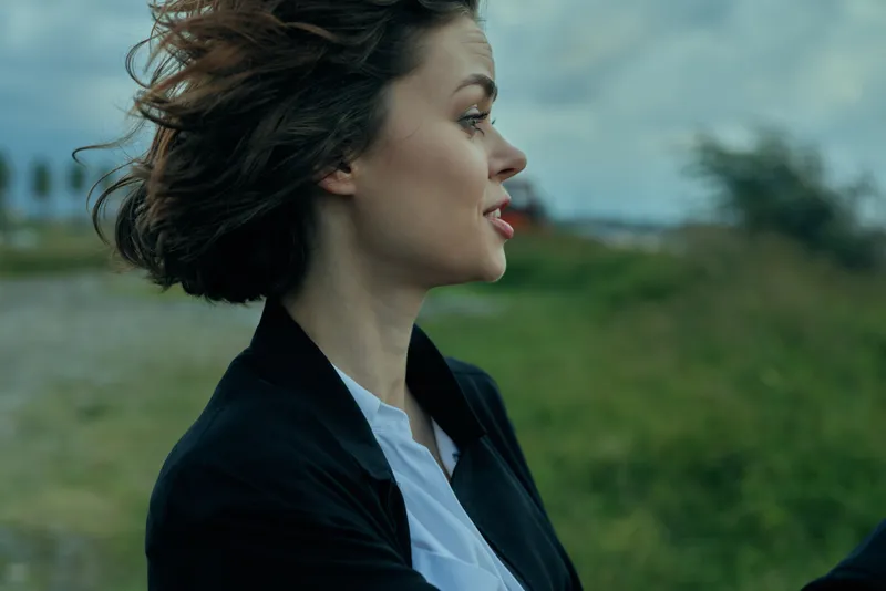 Woman in blazer outdoors, wind in hair, profile facing the horizon, conveying professional focus, calm confidence and determination in a green field.