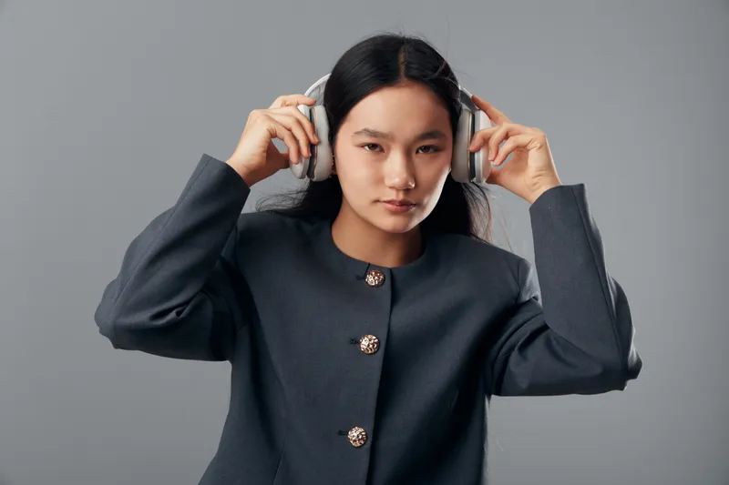 Young woman wearing headphones and gray suit jacket, adjusting audio equipment, portrait with serious expression on gray background. Fashion style and music technology concept.