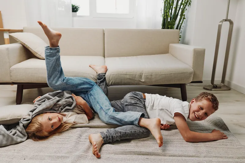Two playful children lying on a carpet in a cozy living room with a beige sofa and natural light. Joyful kids having fun together in a casual home environment. Concept of childhood and happiness.