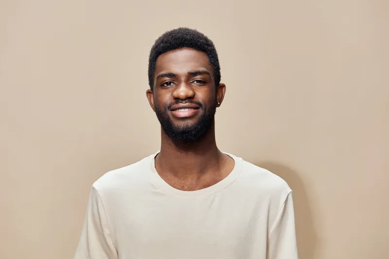 Smiling young African American man with a beard wearing a beige shirt against a neutral background, embodying positivity and confidence in a casual setting