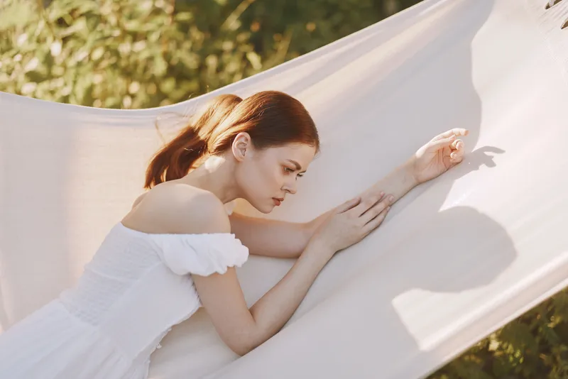 Young woman in a white dress relaxing on a hammock during a sunny day, surrounded by greenery, evoking feelings of tranquility and leisure