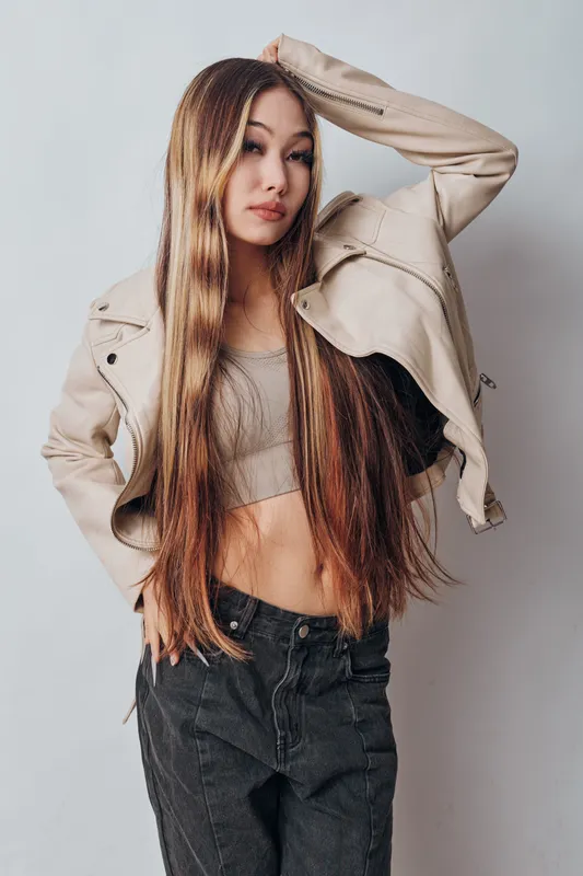 Young woman with long hair wearing beige jacket and crop top posing against white background. Fashion model, casual outfit, trendy style with confident expression.