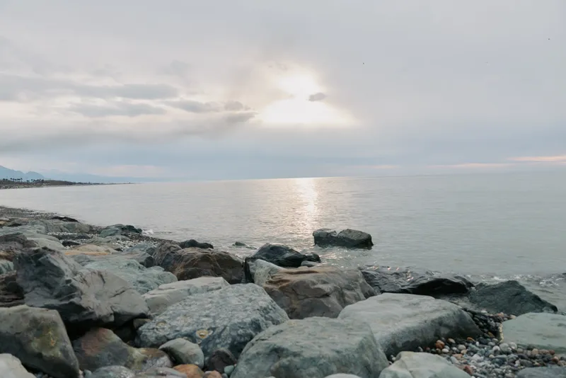 Rocky coastline with calm sea under cloudy sky during sunset. Soft light reflects on water surface creating peaceful and serene atmosphere at the beach shore.