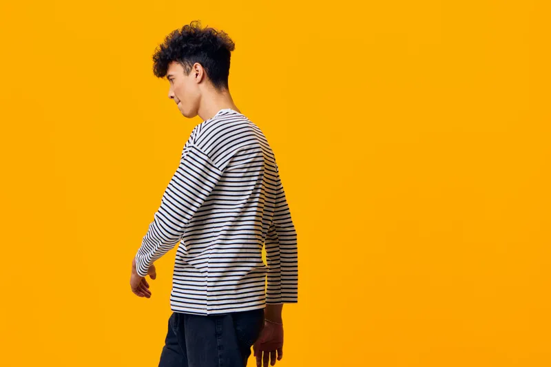young man in striped long sleeve shirt posing sideways with serious expression against vibrant orange background, casual style, contemporary fashion portrait