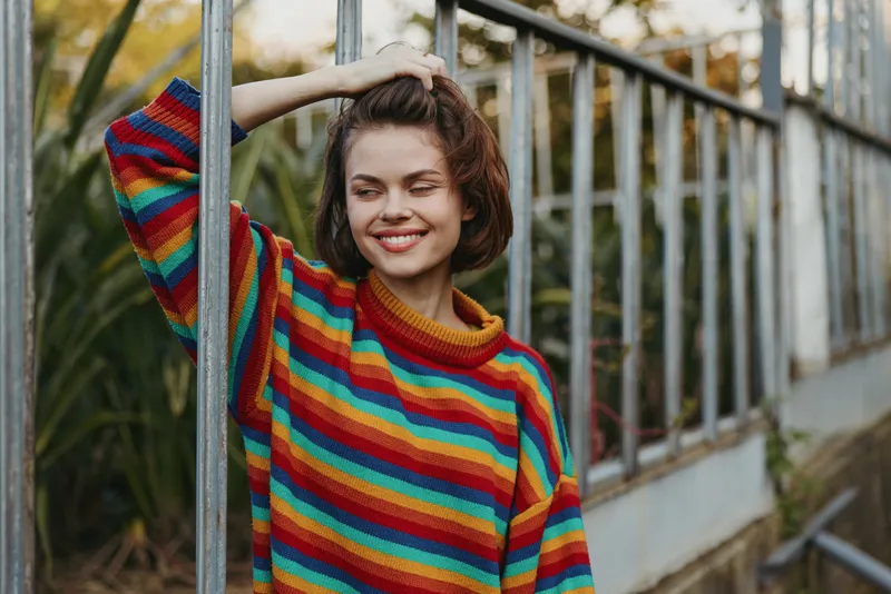 Woman smile rainbow sweater wink portrait in greenhouse, casual young female leaning on metal frame, cozy colorful knit, relaxed outdoor fashion and natural mood.