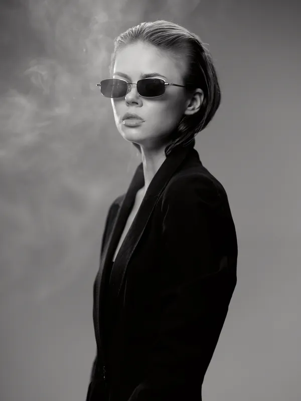 Stylish woman wearing sunglasses and black coat in black and white portrait with smoky background.