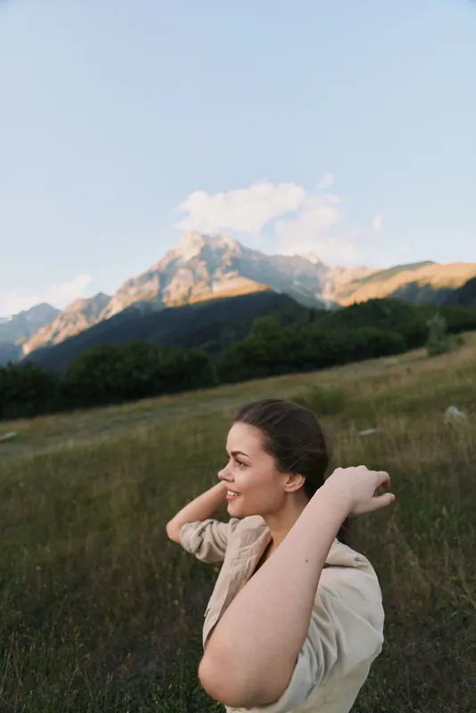 A woman in a light jacket stands in a grassy field with distant mountains, smiling as sunlight warms the landscape. A candid outdoor moment highlighting lifestyle and nature.