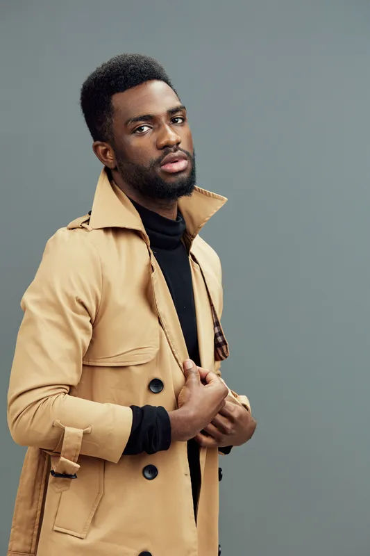 Stylish young man in a beige trench coat against a gray backdrop, showcasing contemporary fashion and a confident expression