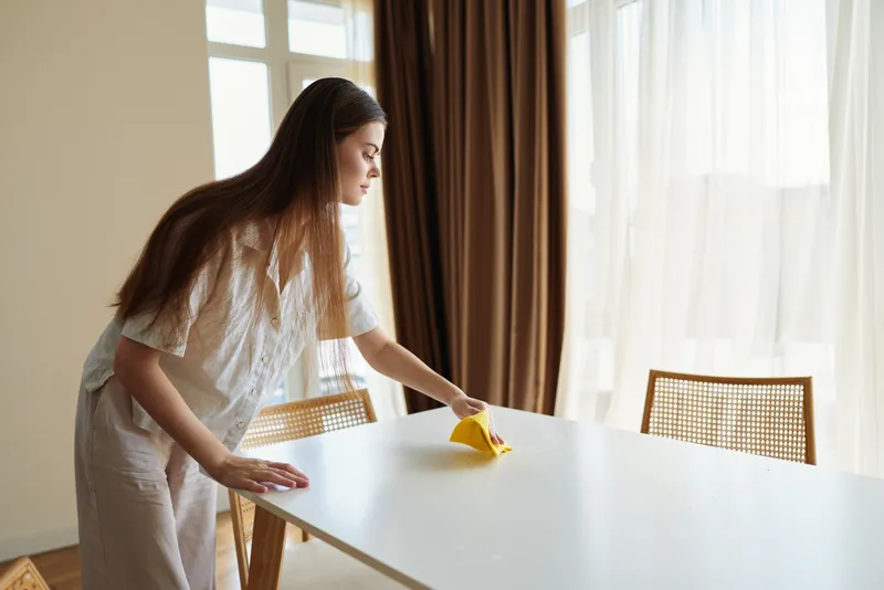 Woman cleaning table in bright dining room, wiping surface with yellow cloth by window. Home chore with sunlight through curtains, wooden chair and tidy minimalist interior for daily upkeep.