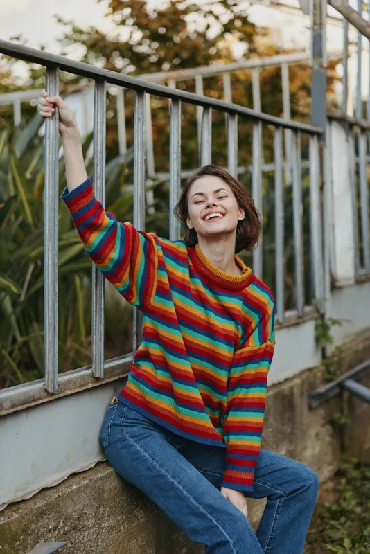 Young woman, striped sweater, smiling, leaning, outdoor, casual outfit: young woman in a colorful striped sweater smiling and leaning on a metal railing in an urban garden, relaxed lifestyle portrait.