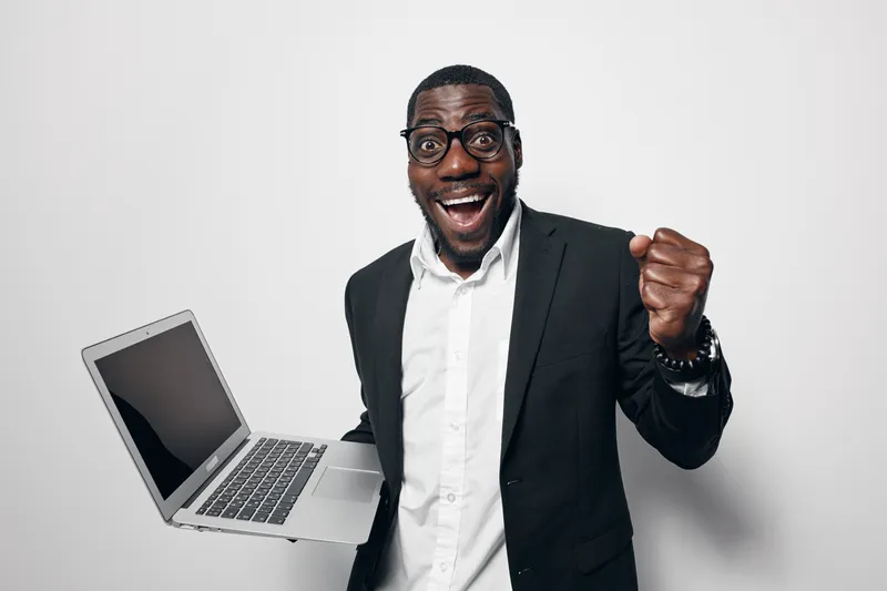African American man with dark skin wearing glasses and suit holding laptop celebrating success with excited expression and clenched fist on white background.