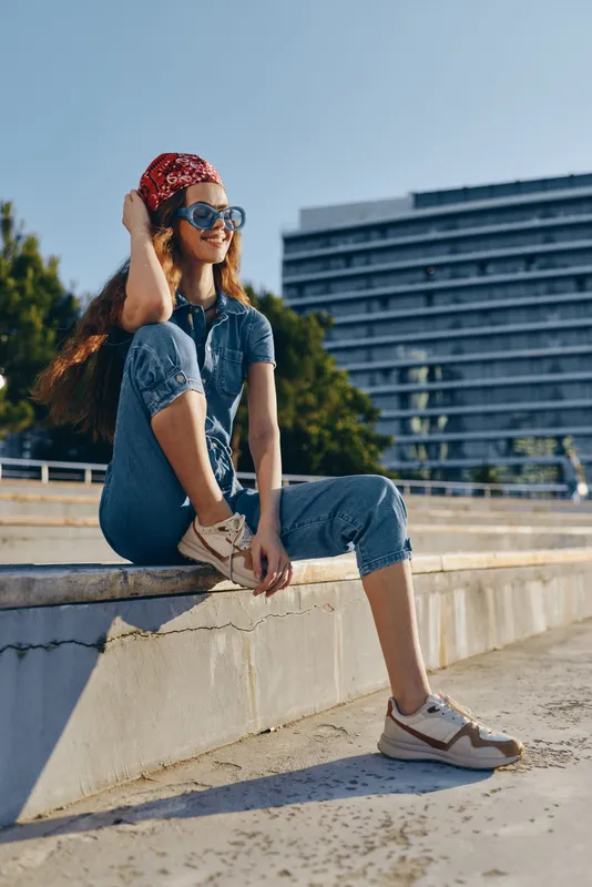 Lifestyle woman in modern boho-western denim street style wearing sunglasses and bandana, sitting outdoors with film color effect and relaxed urban vibe.