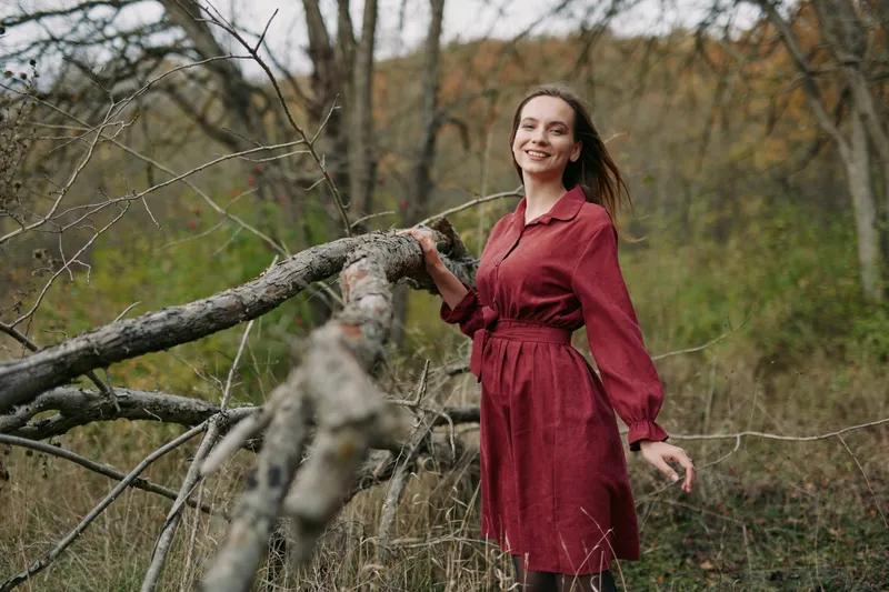 A candid portrait of a woman in a burgundy dress standing beside a fallen tree in an autumn field, showcasing authentic mood and natural lighting.