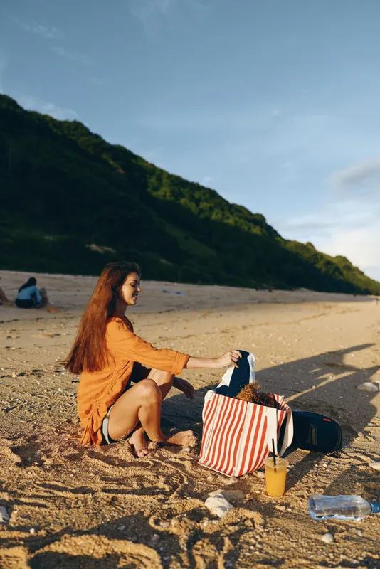 Young woman relaxing on sandy beach in sunny summer light, sitting beside picnic, striped tote bag open, travel backpack and bottle on sand, mountains and blue sky background, calm mood
