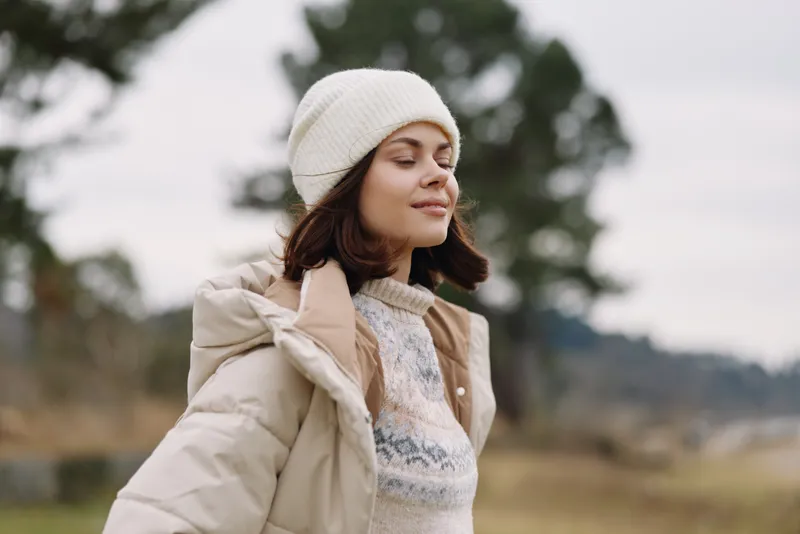 Woman outdoors in a light beige coat and knit sweater, wearing a white beanie, enjoying a breezy moment in a muted landscape, with soft focus and a calm, pensive expression