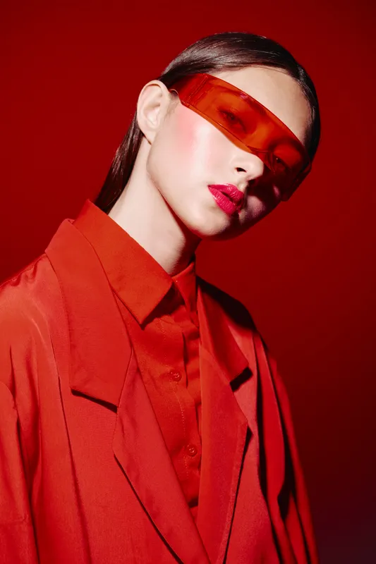 Young woman styled in bold red fashion and vibrant makeup, wearing orange sunglasses in a creative beauty portrait. Studio shot with dramatic lighting and color contrast.