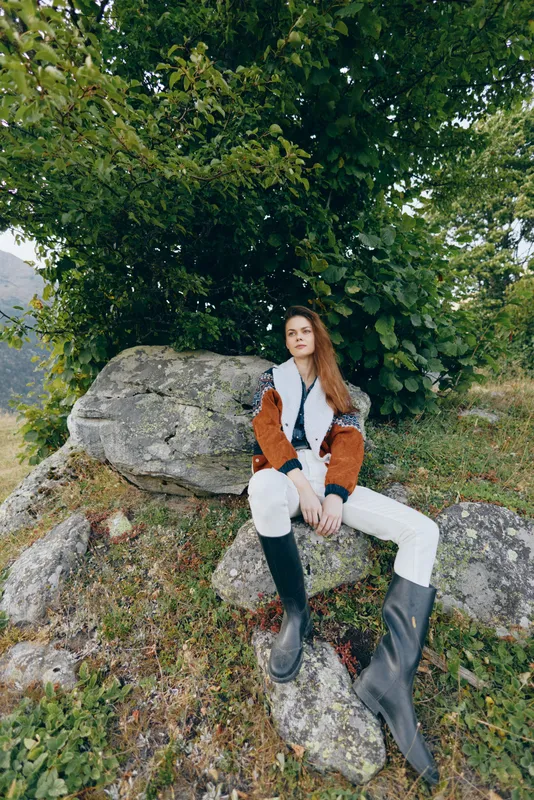 Woman sitting on rock in nature under a tree wearing a brown jacket and tall black boots, outdoor portrait of a relaxed model on a grassy hillside with casual fashion and serene mood.