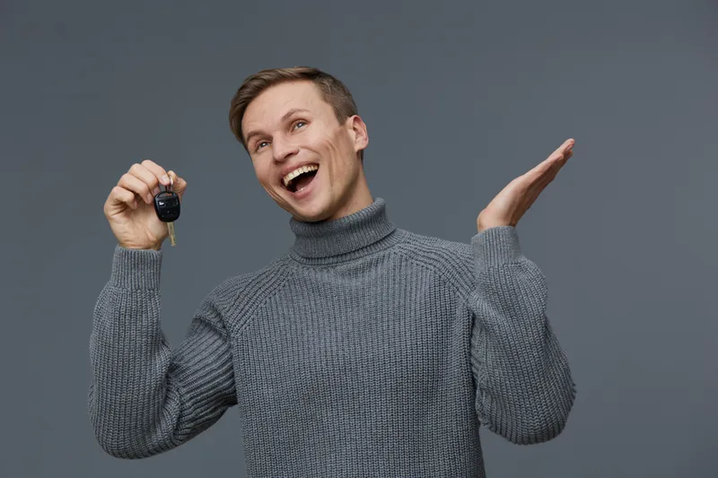 Cheerful young man with short brown hair wearing a cozy gray turtleneck sweater holding key in hand, smiling widely, looking excited, standing against plain gray background. Studio portrait, happy lif
