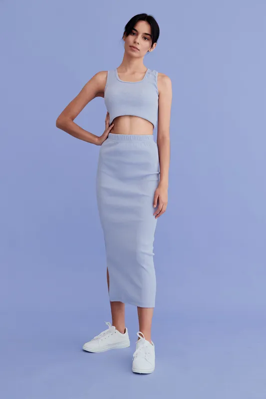 slender woman wearing a light blue crop top and long skirt, posing against a pastel purple background with casual white sneakers for modern minimalistic style