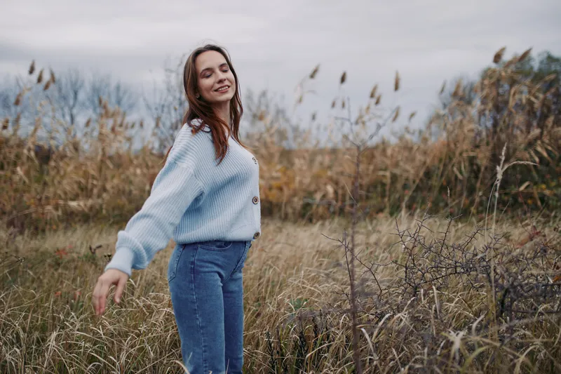 A young woman in a cozy sweater spins gently in a windswept grassy field, captured in a candid pose conveying genuine emotion, authenticity and credibility in a relaxed outdoor lifestyle scene.